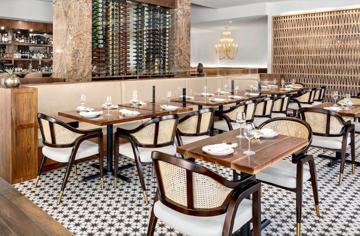 Industrial style restaurant with wooden tables and cane back dining chairs, featuring elegant glassware and table settings.