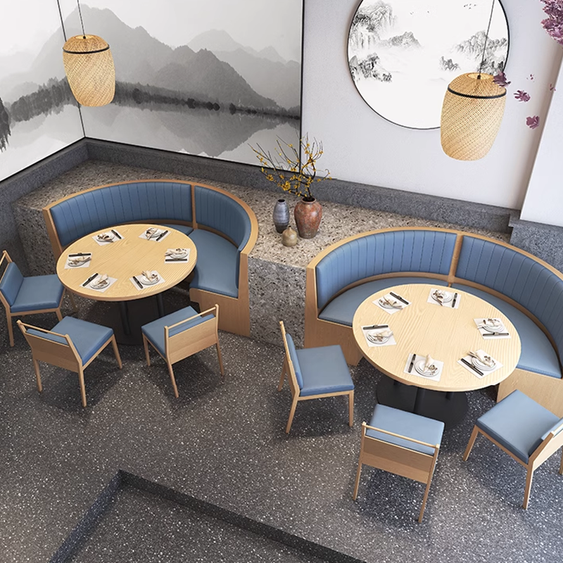 Interior of a restaurant with corner booth seating and light wood tables. The seating has blue leather upholstery, and the room features hanging light fixtures and decorative plants.